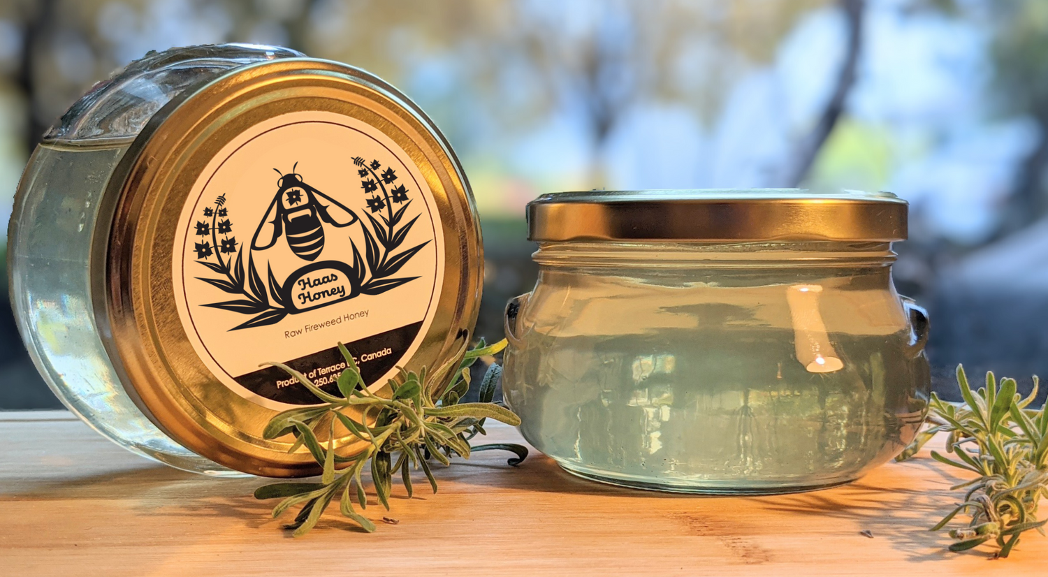 Two jars of honey that are pale yellow in colour. One jar is on it's side and has a black and white logo of a west coast indigenous style honey bee and fireweed plant. Text on the label reads: Haas Honey, Raw Fireweed Honey. Terrace BC 250-635-9020